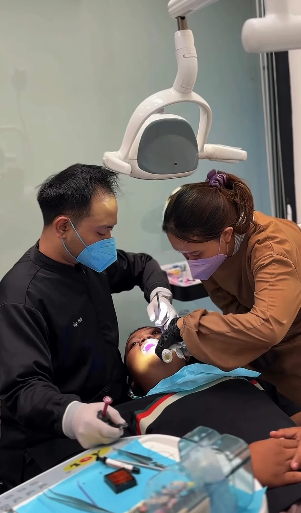 Dokter gigi dan asisten sedang melakukan tindakan di bawah lampu dental